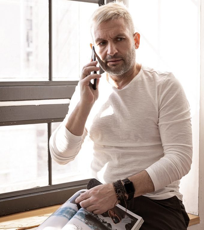 Charming man in a white shirt is sitting on a windowsill with a journal talking on the phone